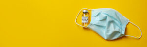 Vial of Covid – 19 vaccine and mask on yellow background