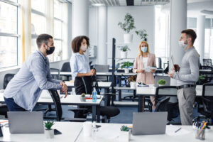Modern team meeting, group work and social distancing. Manager with tablet speaks with workers in protective masks in interior of modern office with gadgets